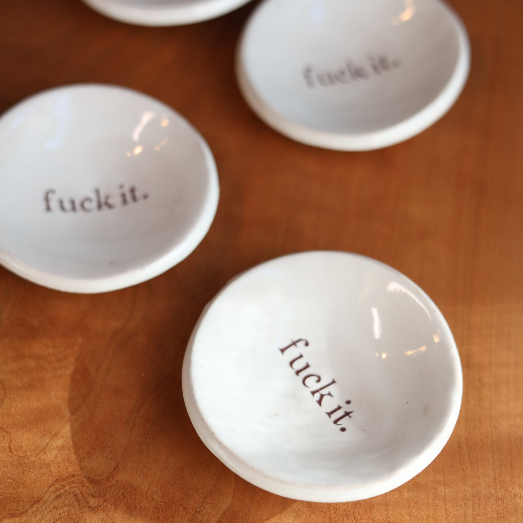 Four white ceramic bowls with 'fuck it.' text on a wooden surface