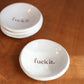 Two white ceramic dishes with 'fuck it.' text on a wooden surface